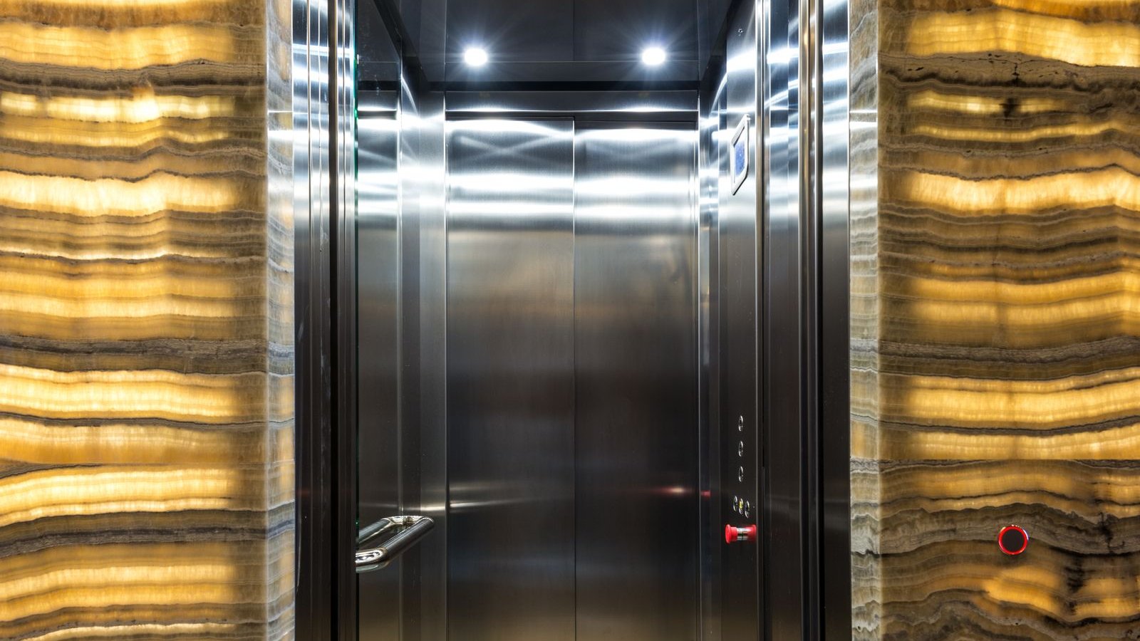 Stainless-steel elevator cab interior framed by backlit stone-veneer panels in a building lobby