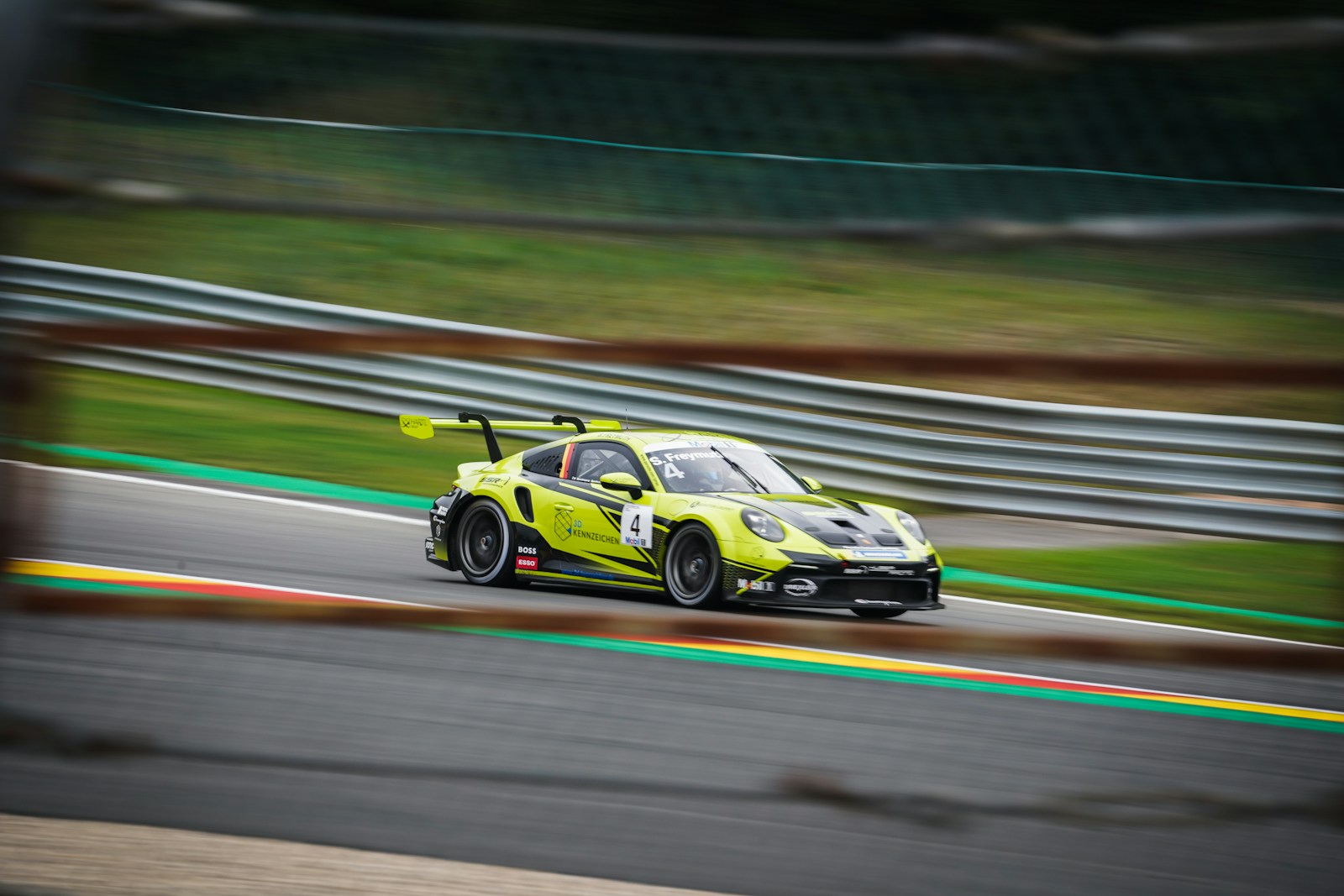Fluorescent yellow-green Porsche 911 GT3 Cup race car cornering on a circuit, panning shot with motion-blurred guardrails.