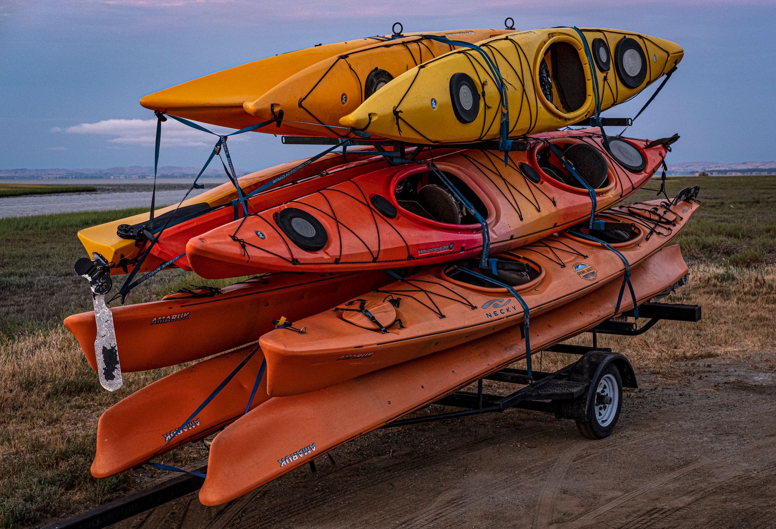 Stack of yellow, red and orange plastic kayaks strapped to a trailer at dusk near the water.