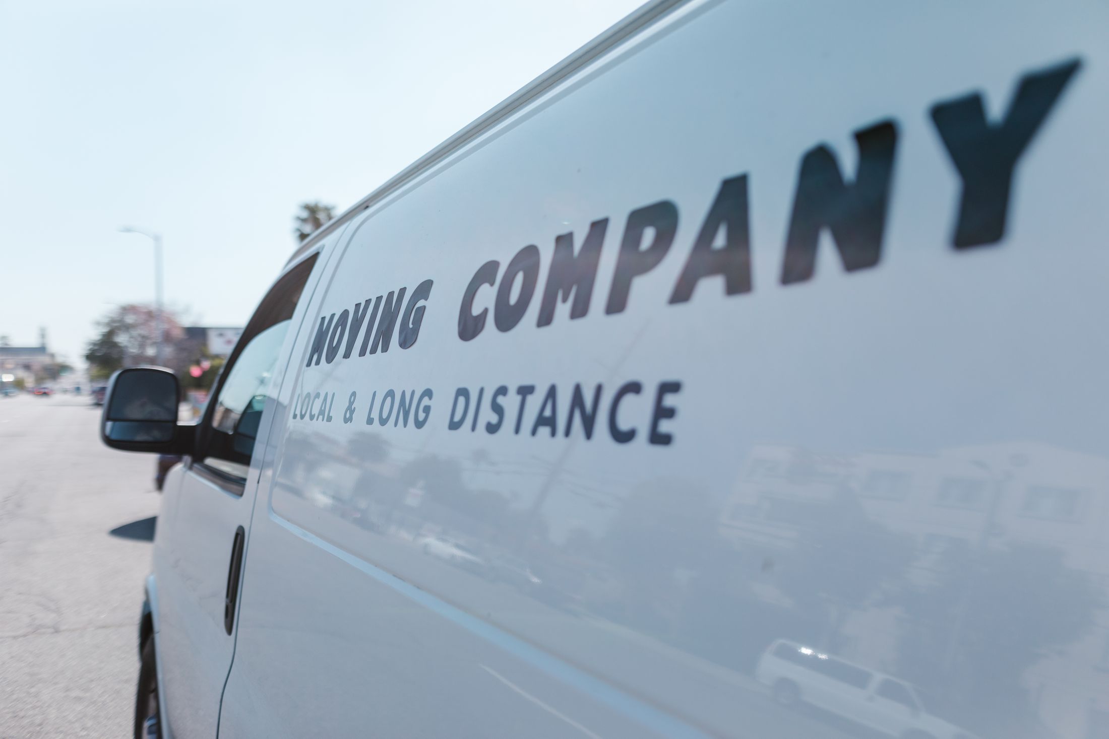 White work van with generic 'Moving Company — Local & Long Distance' lettering on its side panel, parked on a sunny street.