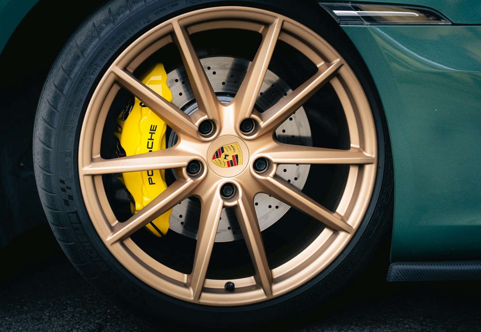 Close-up of a green Porsche's gold ten-spoke wheel with a bright yellow Porsche brake caliper and drilled rotor visible behind the spokes.