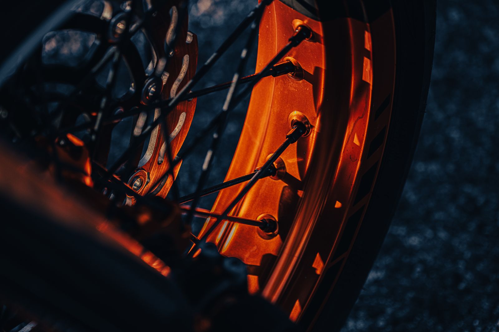 Dusk close-up of a spoked motorcycle wheel, its orange rim glowing in low light with black graphics banding the rim lip.