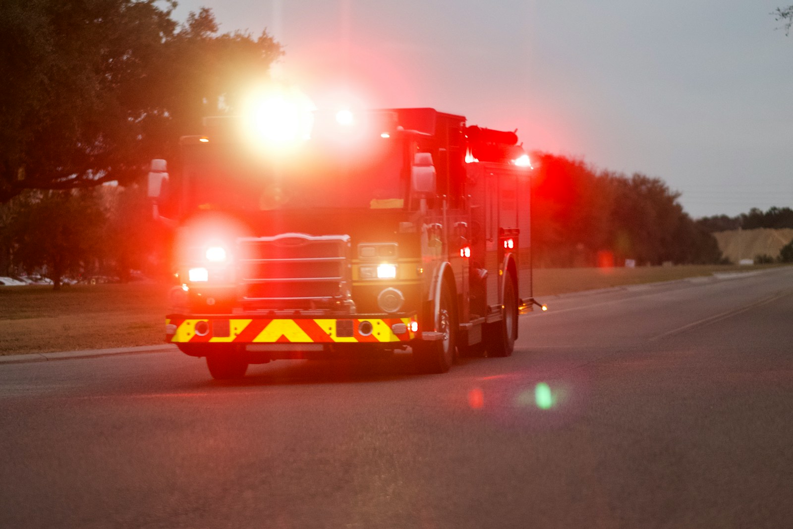 Fire engine on a road at dusk with emergency lights glowing and a retroreflective red-and-yellow chevron panel across its front bumper.