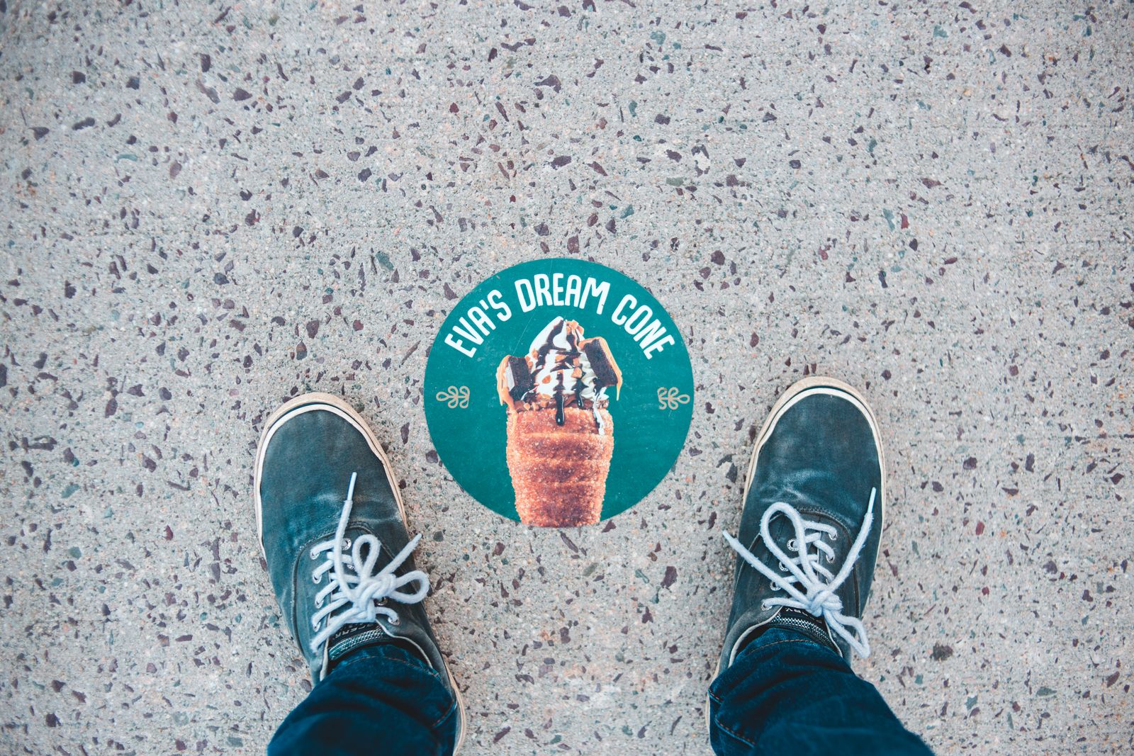 Round printed ice-cream-shop decal applied to a concrete sidewalk, seen from above with sneakers standing beside it