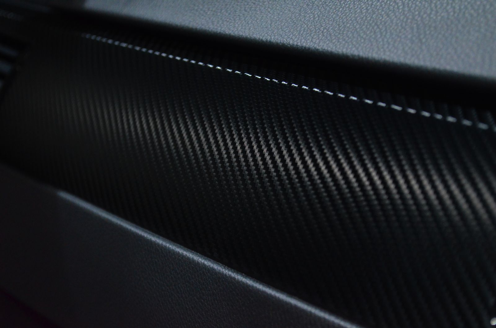 Close-up of carbon-fiber weave texture on a car's trim panel, bordered by black leather grain with white stitching.