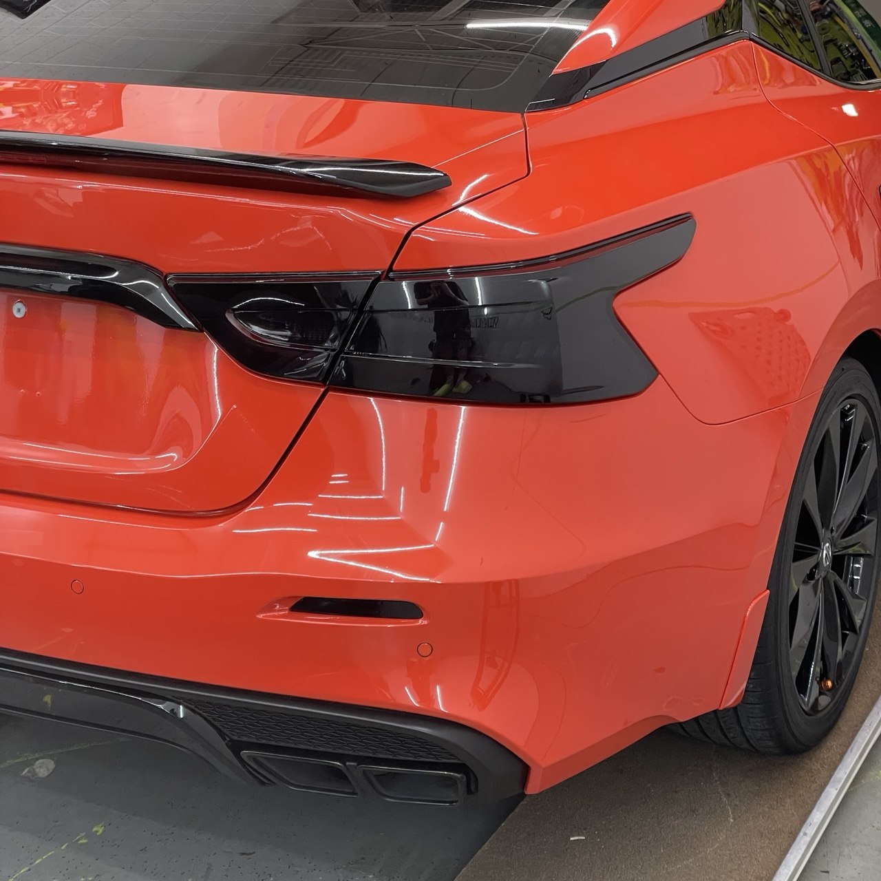 Rear three-quarter of an orange sedan in the shop with smoked-out taillights and gloss-black spoiler, badges and trim.
