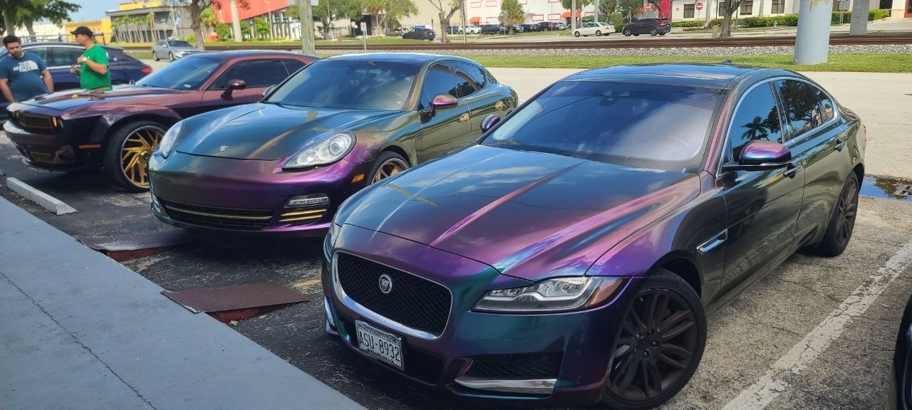 Three color-shift wrapped cars in daylight — an iridescent purple-teal Jaguar XF in front, a purple-green Porsche Panamera and a dark color-shift muscle car behind it, each showing two colors in the same frame.